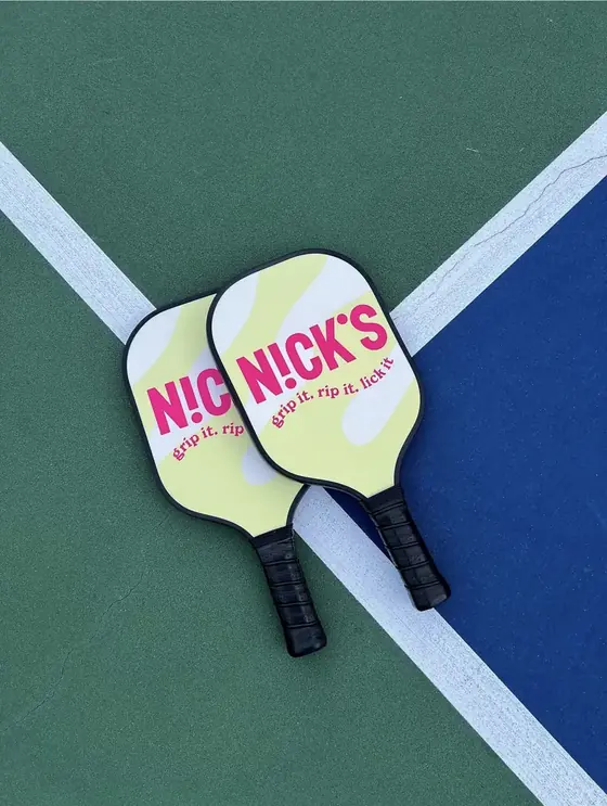 Two pastel yellow Nick's branded pickleball paddles on a court line
