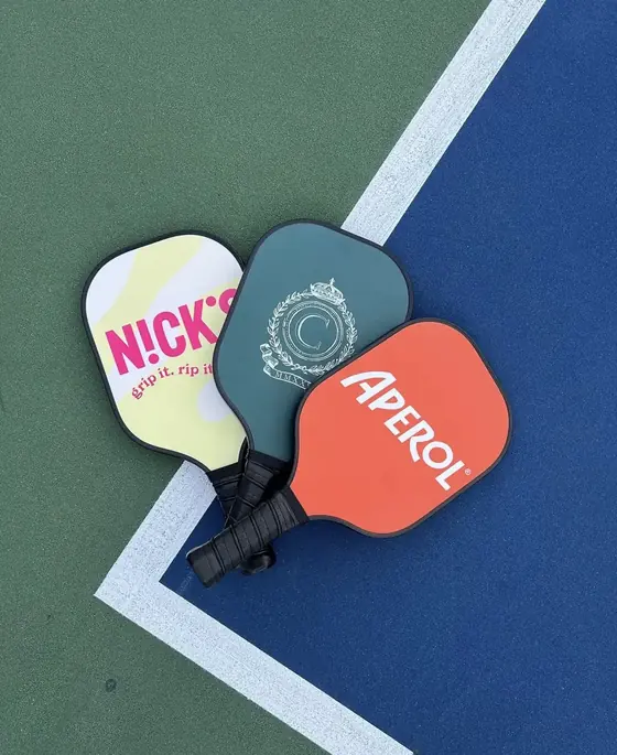 Three custom pickleball paddles including an orange Aperol paddle on a court